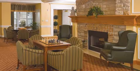 A cozy living room area in Breckenridge Village featuring a stone fireplace with a wooden mantle, two dark green armchairs, a wooden table with a chess set, and several striped upholstered chairs. The room has warm yellow walls, carpeted flooring, and a window with curtains letting in natural light.