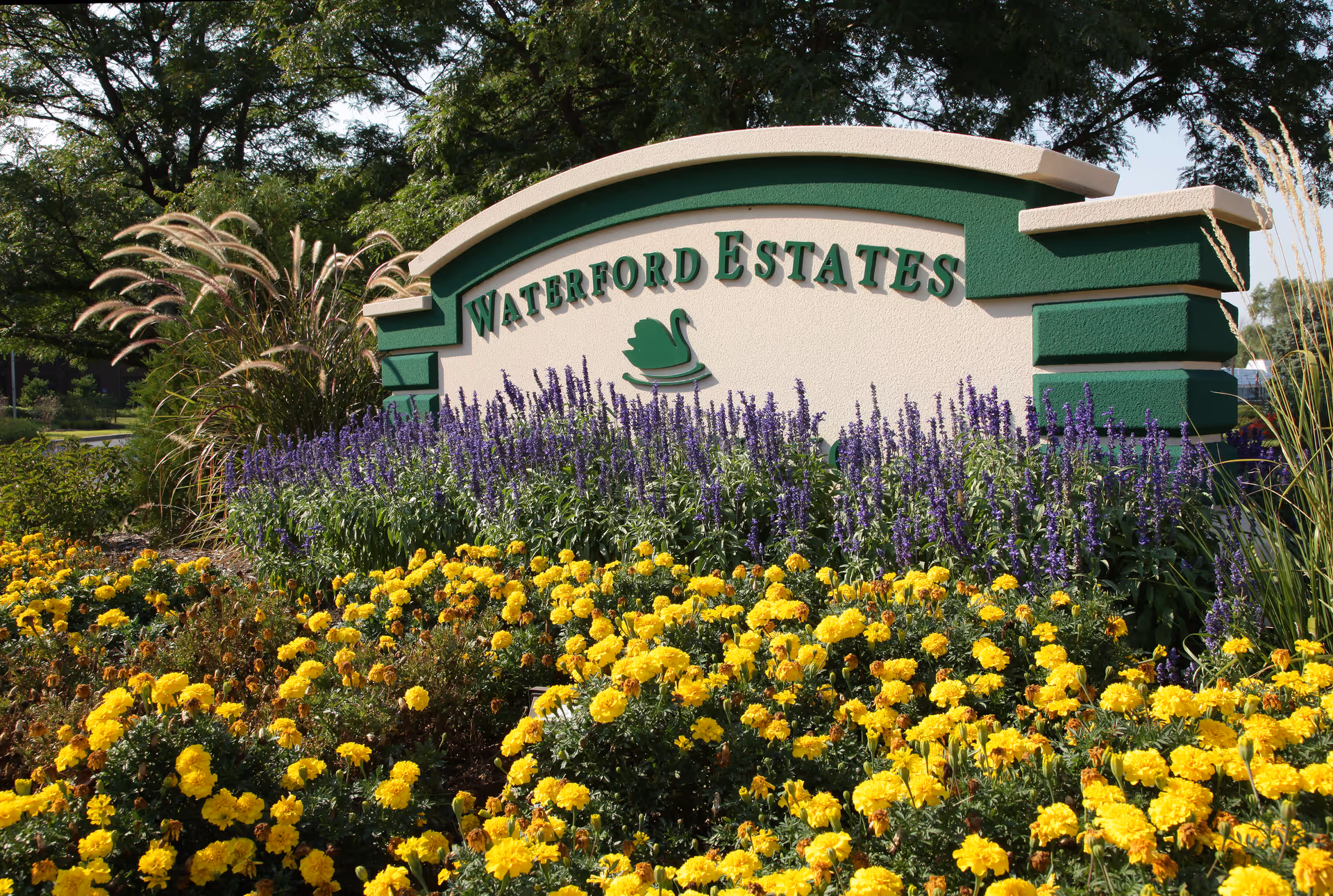 Entrance sign reading "Waterford Estates" with a swan logo surrounded by purple and yellow flowers and landscaping.