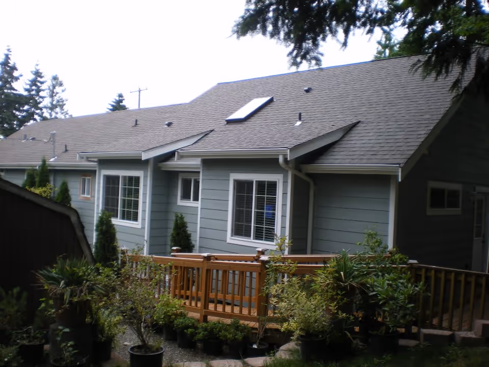Exterior view of a single-story residential building with gray siding and a shingled roof, featuring multiple windows and a wooden deck surrounded by various potted plants and shrubs.