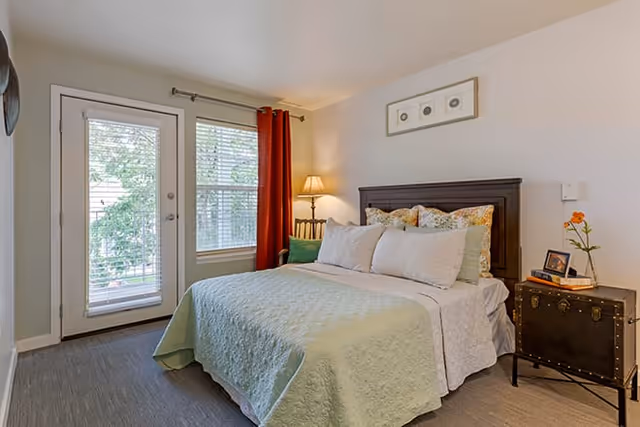 A cozy bedroom with a neatly made bed featuring a light green quilt and multiple pillows. There is a wooden headboard, a bedside table with a lamp, a small chest with a framed photo and a flower vase, and a door and window with blinds and red curtains letting in natural light.