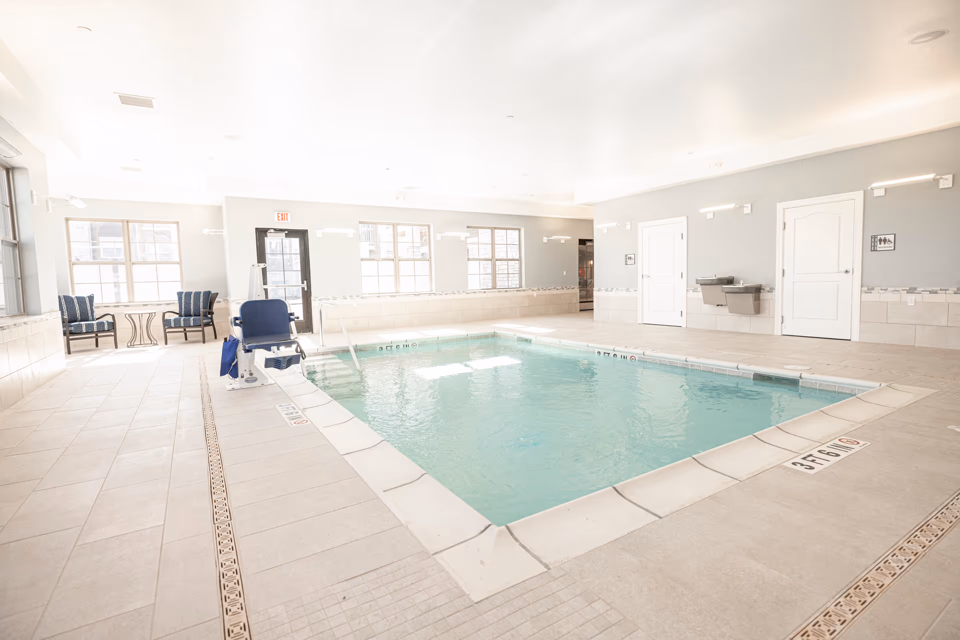 Bright indoor swimming pool with a pool lift, seating, and large windows in a senior living facility.