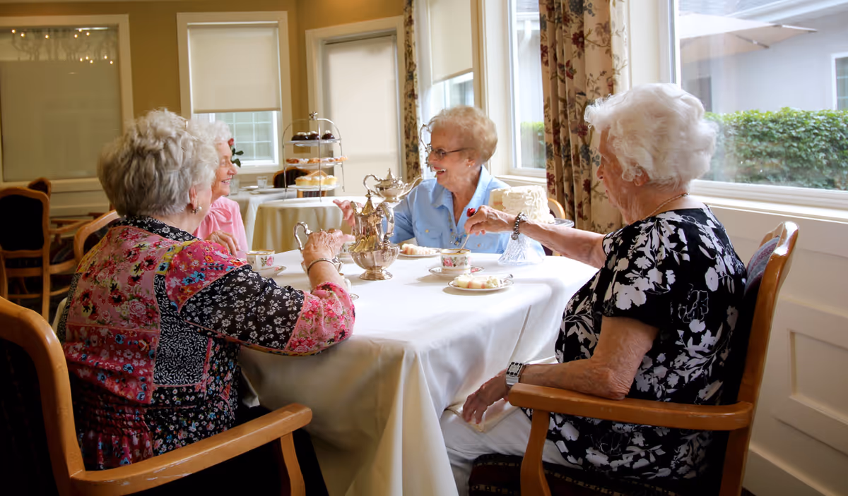 Four elderly women sitting around a table in a bright dining room, enjoying tea and cake. The table is covered with a white tablecloth and has a silver tea set and plates with slices of cake. Large windows with floral curtains let in natural light.