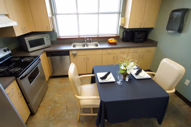 A small kitchen with stainless-steel stove and dishwasher, light wood cabinets, a sink under a window, and a small table set for two.