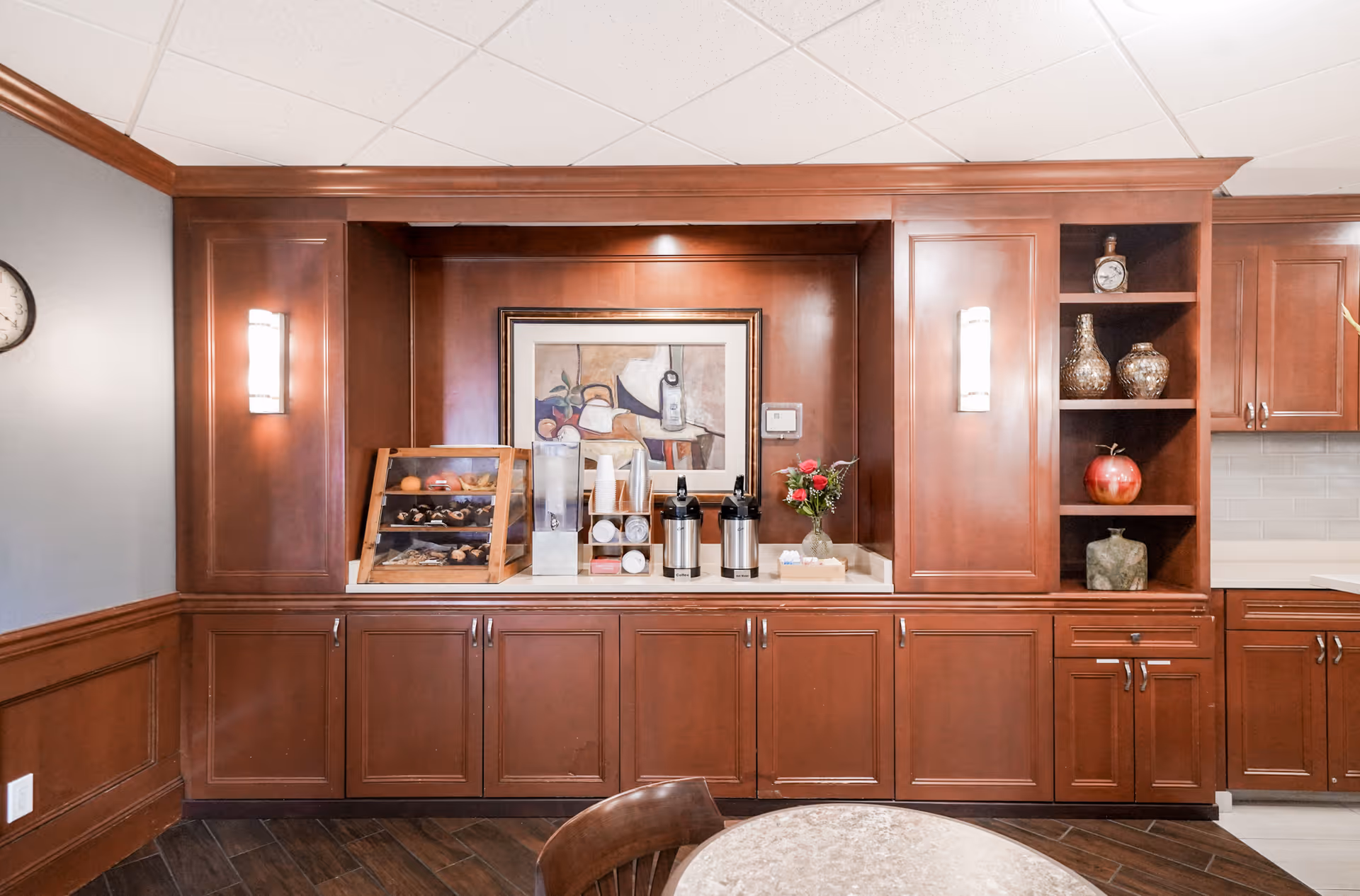 A coffee and snack station set within a wooden built-in cabinet area featuring dark wood cabinetry, a framed artwork on the back wall, two wall sconces, and decorative vases on open shelves. There are coffee dispensers, cups, and a display case with pastries on the countertop. A round table with chairs is partially visible in the foreground.