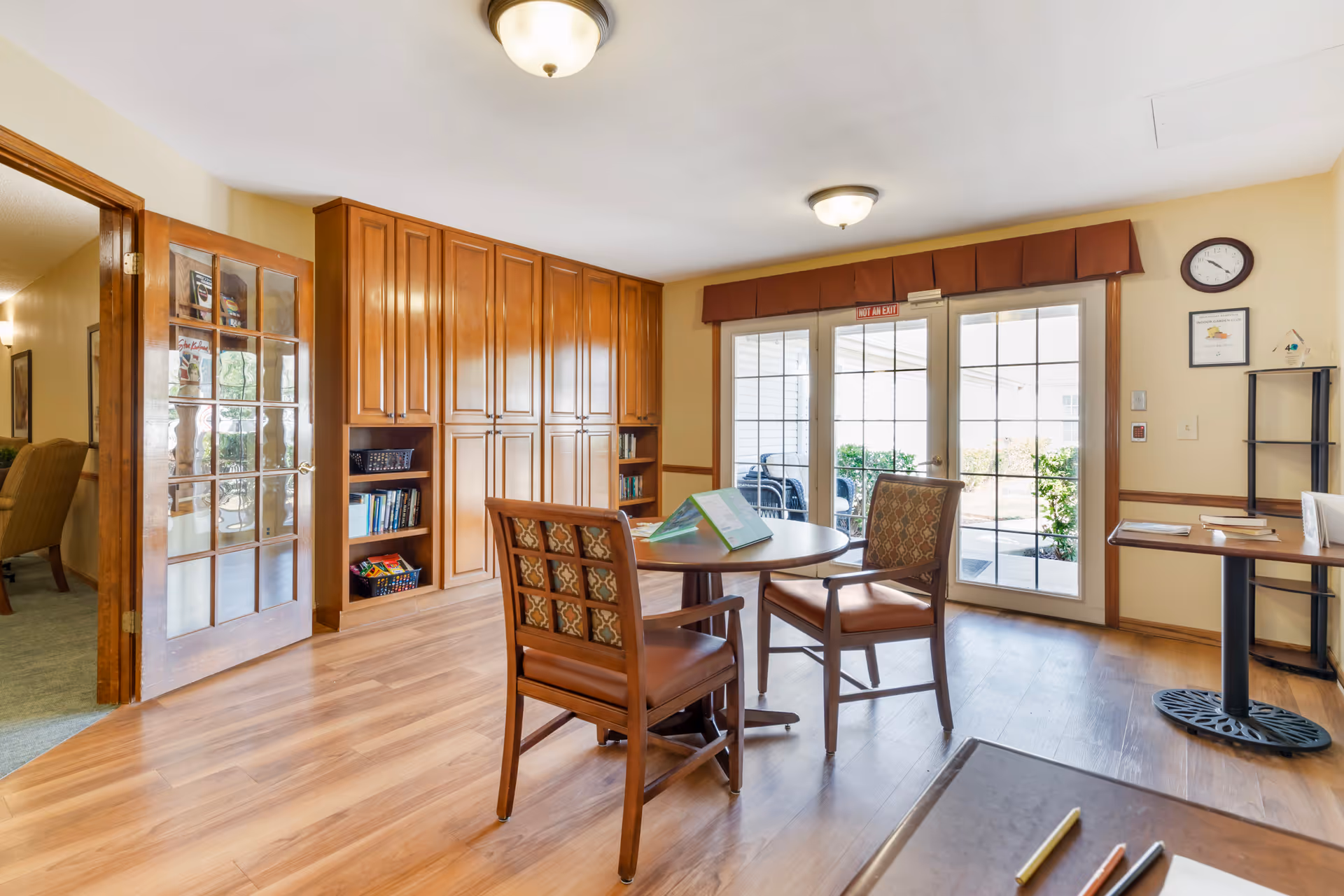 A bright room with wooden flooring and cabinetry, featuring a round table with two cushioned chairs. Glass double doors lead to an outdoor patio area with seating. The room has beige walls, a clock, and a small table with books and papers on the right side.