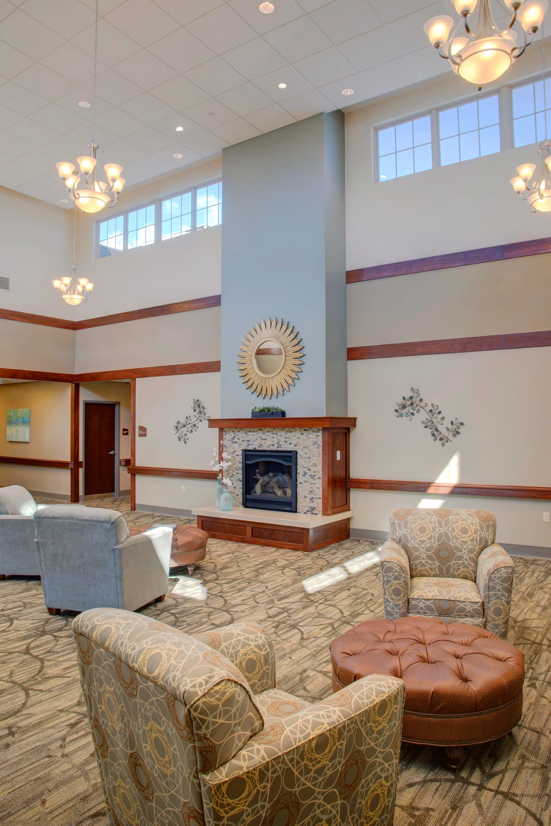 A spacious senior living facility common area with high ceilings and large windows letting in natural light. The room features a central fireplace with a decorative sunburst mirror above it, surrounded by comfortable armchairs and a tufted ottoman. The walls are adorned with simple decorative metal art, and the carpet has a patterned design.