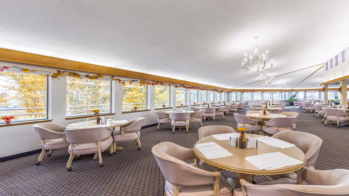 A spacious dining room with multiple round tables and cushioned chairs arranged neatly. The room has large windows along one side, allowing natural light to brighten the space and showing autumn-colored trees outside. The ceiling features elegant chandeliers, and the tables are set with placemats, napkins, and small decorative items.