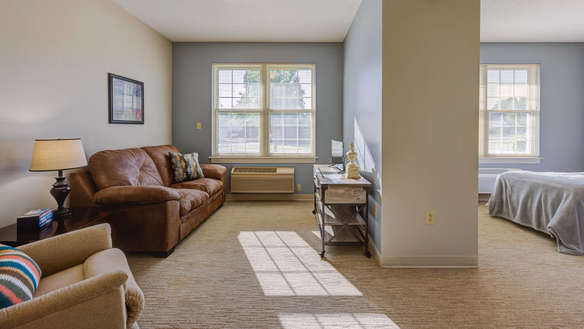 A bright room with beige carpet featuring a brown couch, a beige armchair with a colorful pillow, a side table with a lamp and books, and a window with blinds letting in sunlight. To the right, a partial view of a bedroom with a bed covered in a gray blanket and another window with blinds.