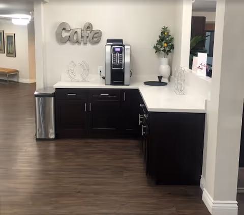 A coffee station area in a senior living facility with a coffee machine on a white countertop, dark wood cabinets below, a metal trash can to the left, and a decorative vase with a small plant on the counter. The word 'Cafe' is mounted on the wall above the coffee machine.