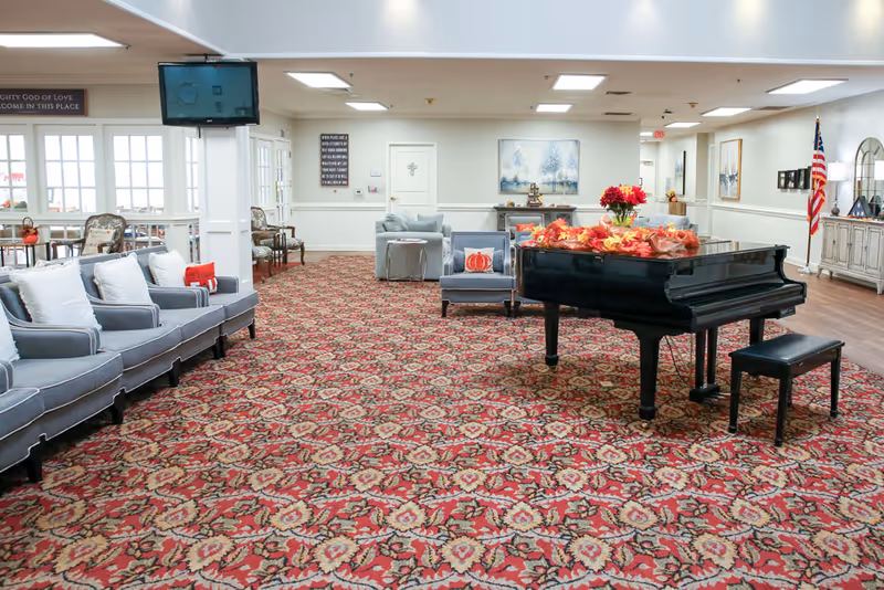A spacious common area in a retirement facility featuring a patterned red carpet, a black grand piano decorated with autumn leaves and flowers, several gray upholstered chairs and sofas with white cushions, and an American flag in the corner. The room has white walls, ceiling lights, and framed artwork on the walls.