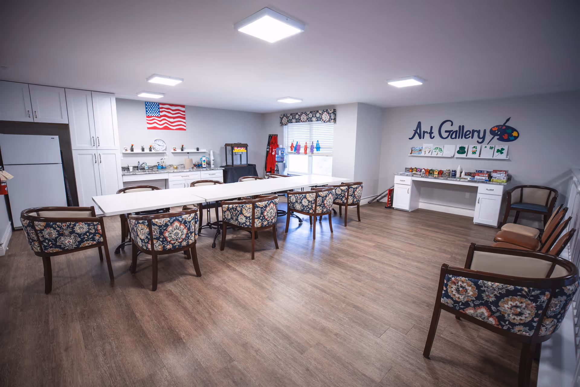 A spacious activity room with a long table surrounded by floral-patterned chairs. The room features a kitchenette area with a refrigerator, cabinets, and a countertop with a sink. An American flag is displayed on the wall above the kitchenette. On the right side, there is a white desk labeled 'Art Gallery' with various art supplies and small crafts displayed. The room has wood flooring and bright ceiling lights.
