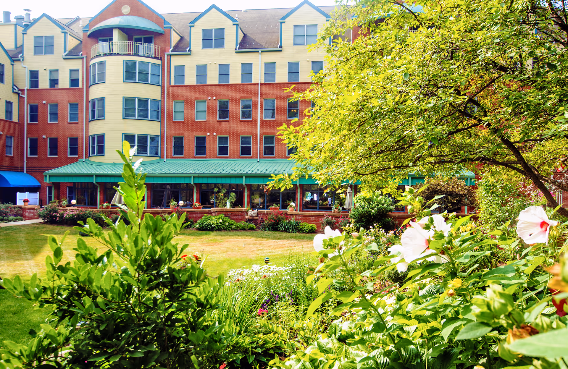 Courtyard garden with flowering plants in front of a multi-story brick and yellow senior living building with green awnings.