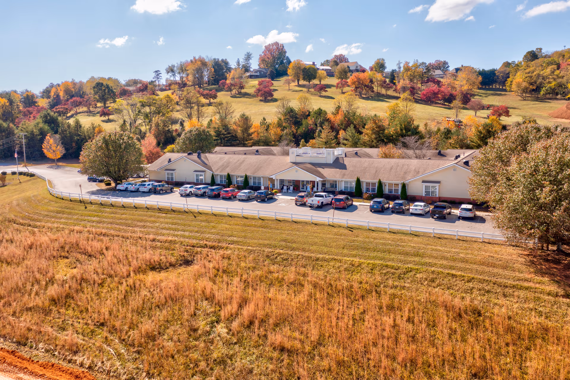 Aerial view of a single-story building with a parking lot filled with cars in front, surrounded by autumn-colored trees and grassy fields under a partly cloudy sky.
