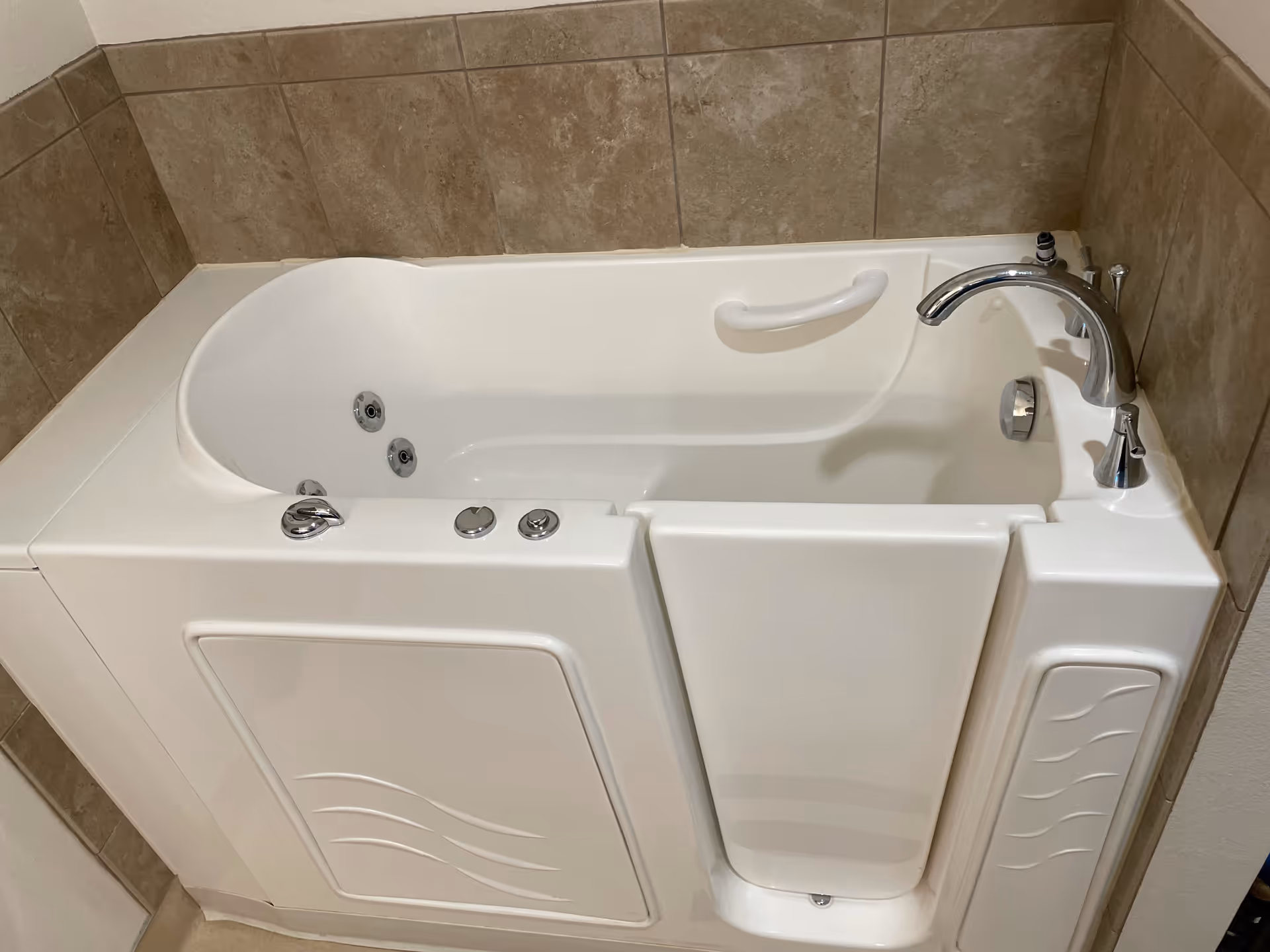 A white walk-in bathtub with a side door and built-in grab bar, surrounded by beige tiled walls, featuring chrome faucet and controls.