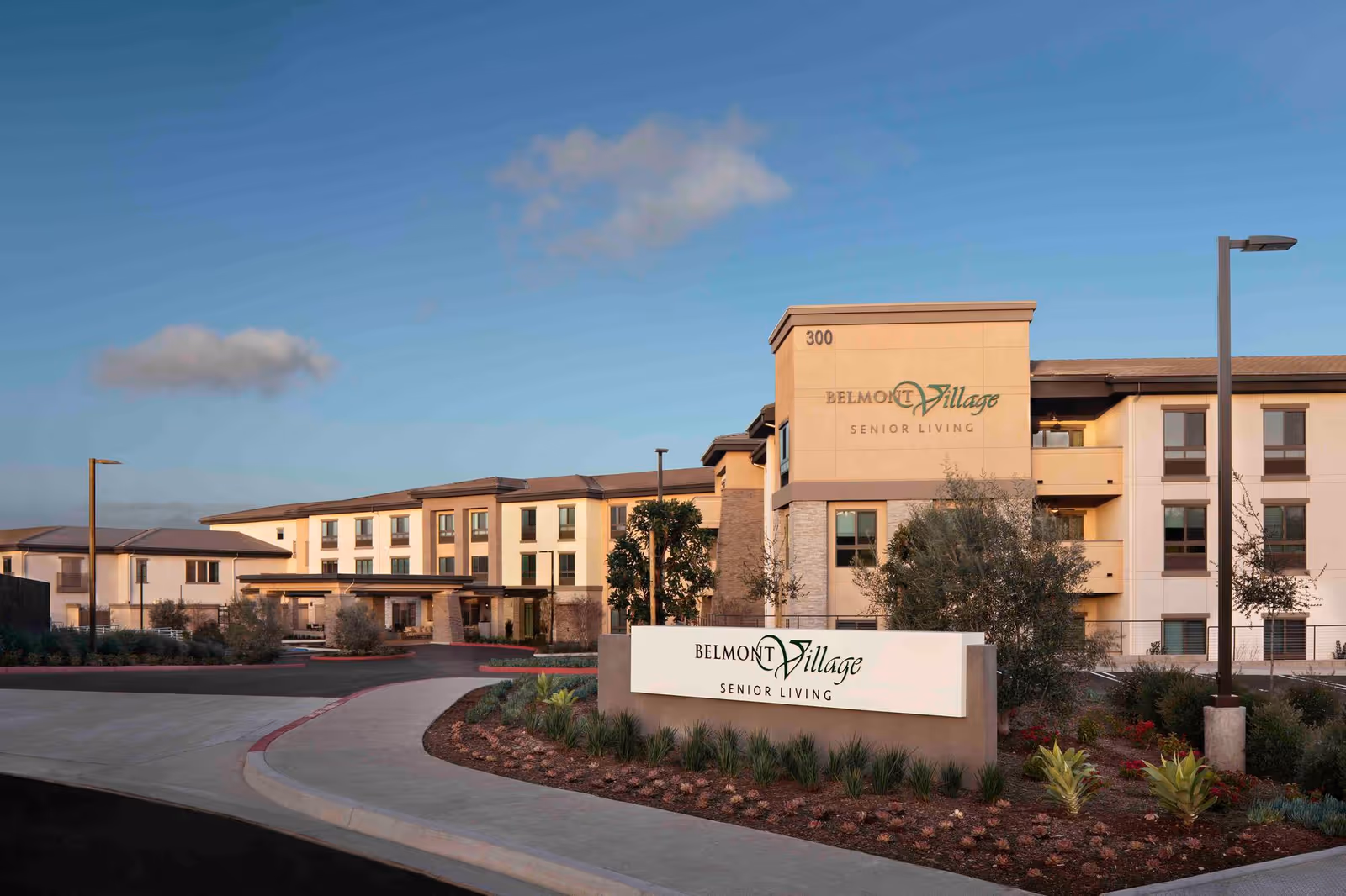 Exterior view of Belmont Village Senior Living Aliso Viejo building with a clear sky, landscaped garden, and a large sign displaying the facility's name in front.