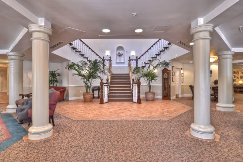 Spacious senior living facility lobby with a central carpeted staircase, decorative columns, potted plants and seating areas.