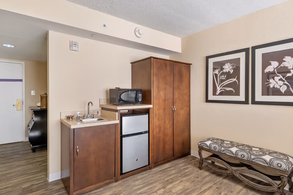 Small kitchenette area with a sink, mini-fridge, microwave and wooden cabinets next to a patterned bench and framed artwork.