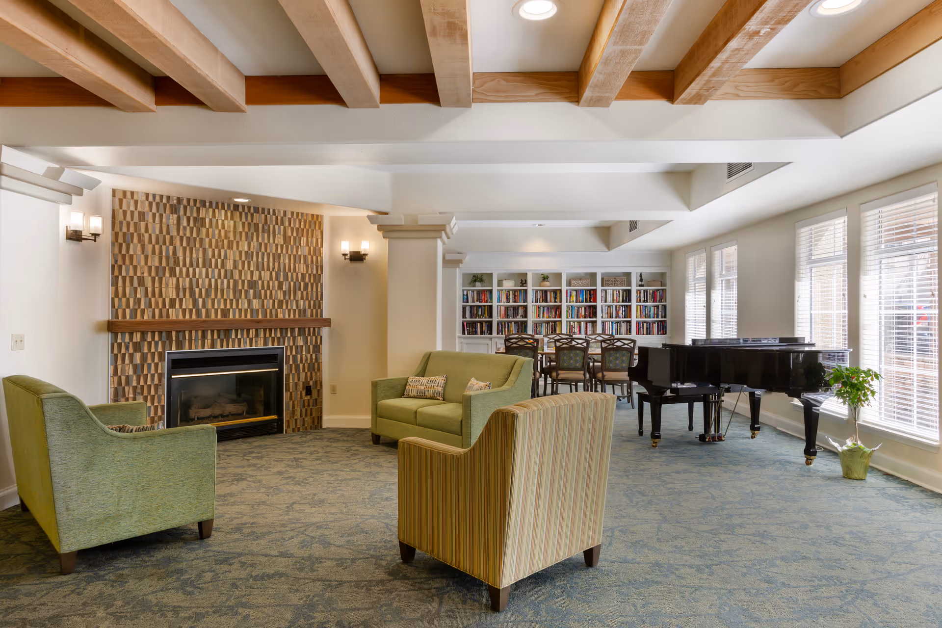 Well-lit communal living room with sofas and armchairs, a fireplace with decorative tile, a bookshelf, and a grand piano under exposed wooden beams.