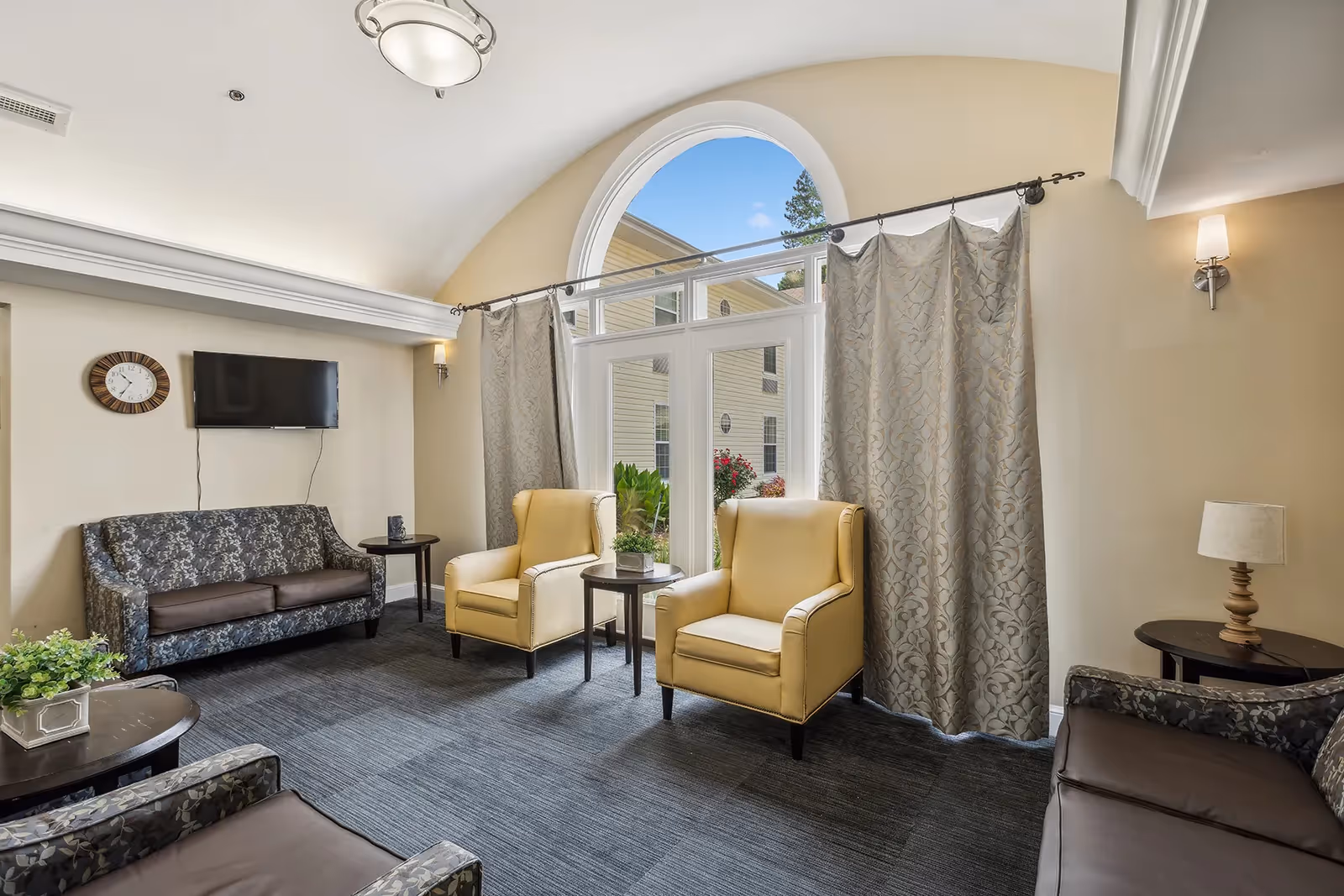 A cozy living room area with two yellow armchairs and two patterned sofas arranged around small round tables. There is a large arched window with curtains, a wall-mounted TV, a clock, and soft lighting fixtures on the beige walls.