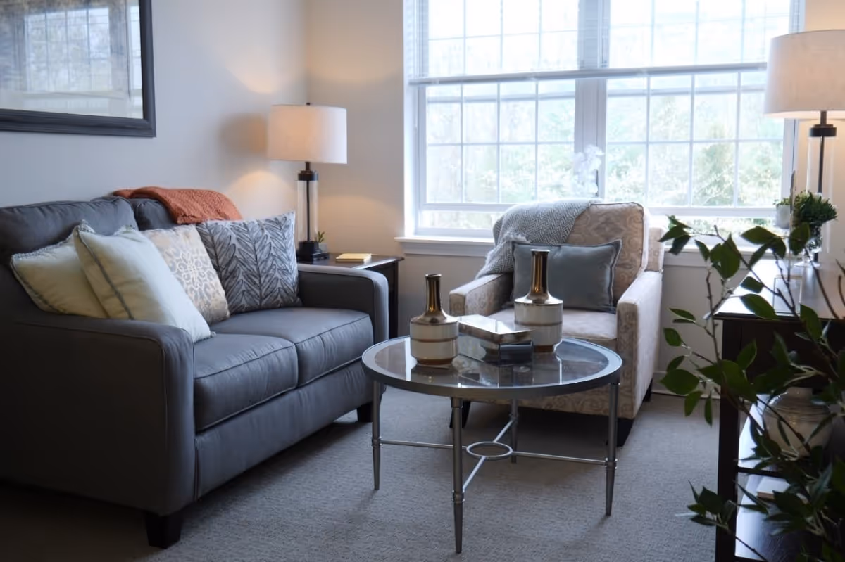 A cozy living room with a gray sofa adorned with multiple decorative pillows, a patterned armchair with a blue pillow and a throw blanket, a round glass coffee table with two decorative vases and a box, two table lamps on side tables, and a large window letting in natural light.