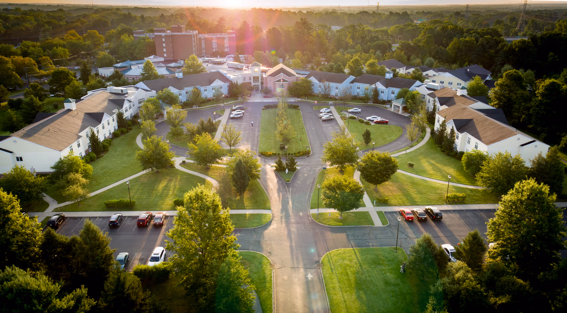 Aerial view of The Terraces at Lawrence senior living facility during sunset, showing multiple white buildings with brown roofs arranged around a central driveway and parking areas, surrounded by green lawns and trees.
