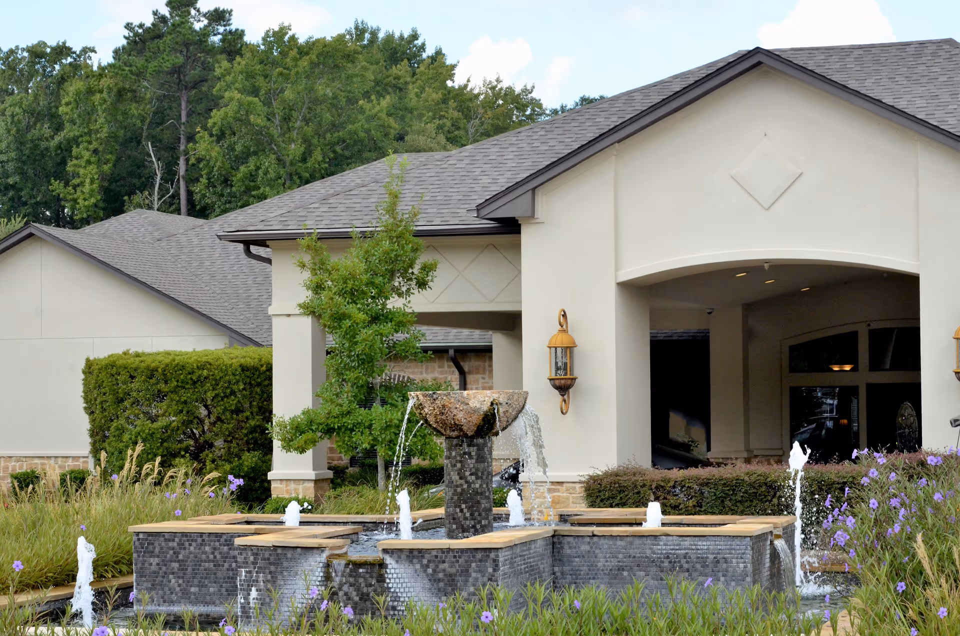 Exterior view of a building with a beige facade and a dark shingled roof, featuring a central covered entrance. In front of the building is a decorative water fountain with multiple streams of water, surrounded by greenery and purple flowers. Trees and bushes are visible in the background.