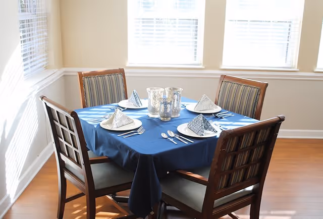 A dining table set for four with a blue tablecloth, white plates, folded napkins, and silverware in a bright room with three large windows and wooden chairs with striped cushions.