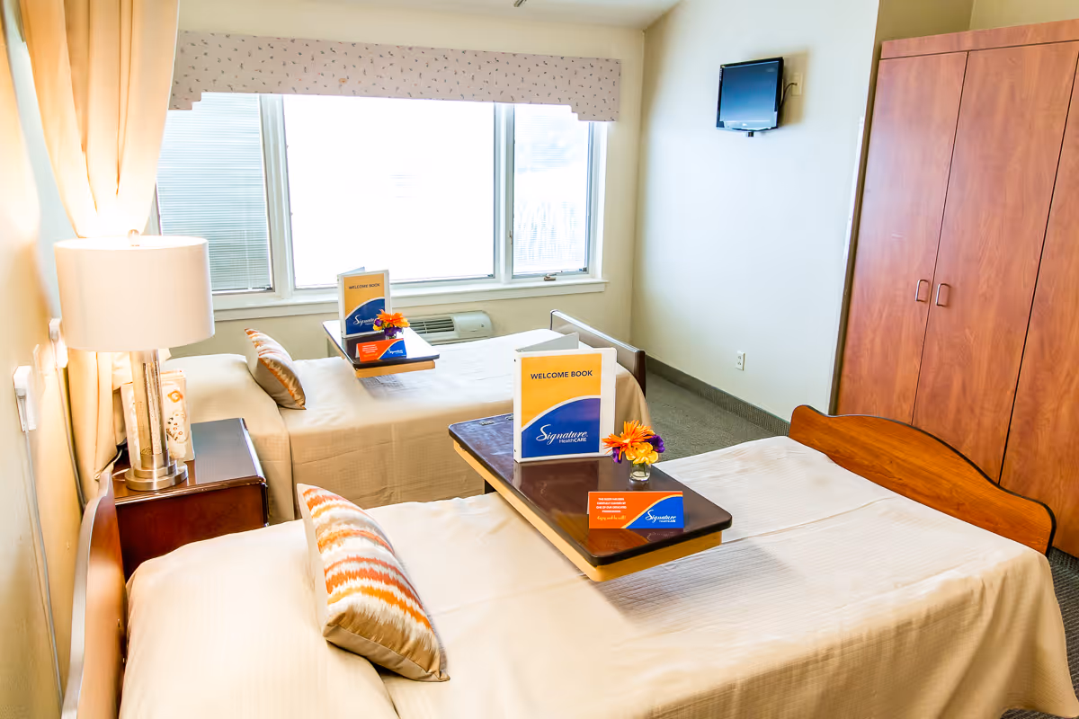 A bright, clean room with two single beds, each with a pillow and a beige bedspread. Each bed has a small overbed table with a welcome book, a small flower arrangement, and a card. There is a wooden nightstand with a lamp between the beds. A large window with blinds and a valance lets in natural light. A wall-mounted TV is visible on the right wall, and a wooden wardrobe is in the corner.