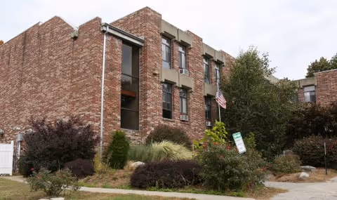 Brick three-story rehabilitation and health care center building with landscaped shrubs and an American flag in front.