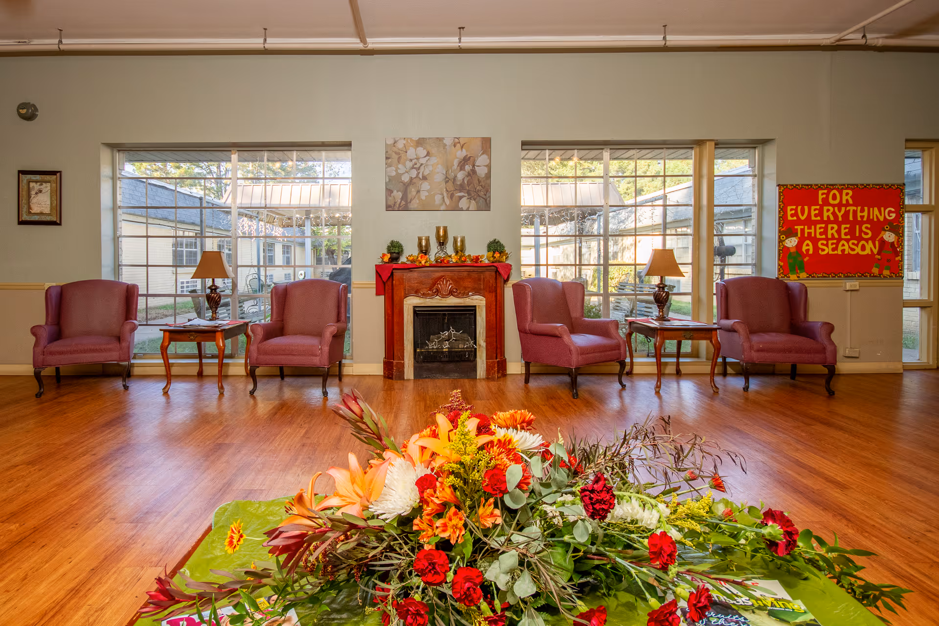 A cozy common area with four purple armchairs arranged around two small wooden tables with lamps. There is a decorative fireplace in the center with fall-themed decorations and a floral painting above it. Large windows behind the chairs provide a view of an outdoor courtyard. A colorful bulletin board on the right wall reads 'For Everything There Is A Season.' A large bouquet of flowers is in the foreground on a table.