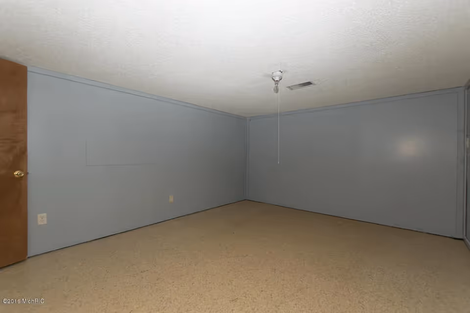 Empty interior room with light blue walls, beige carpet, a wooden door, and a ceiling light fixture with pull cord.