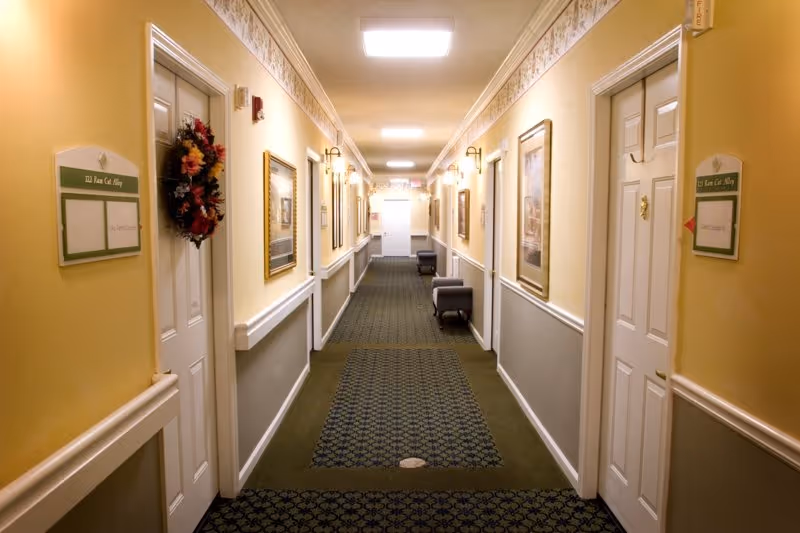 Well-lit carpeted interior hallway in a senior living facility lined with apartment doors, framed artwork, and seating.