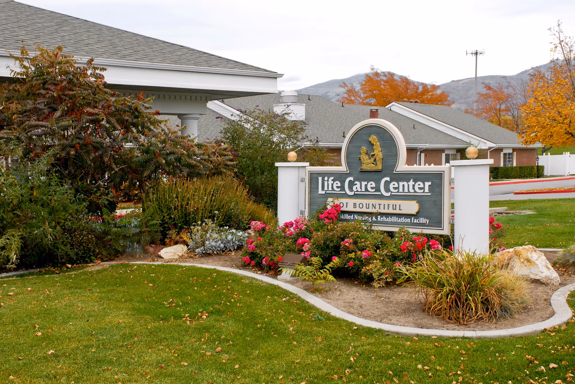 Outdoor view of the entrance area of Life Care Center of Bountiful, a skilled nursing and rehabilitation facility, featuring a landscaped garden with green grass, bushes, and colorful flowers in front of the facility sign. The building and mountains are visible in the background under a cloudy sky.