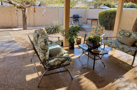 Covered outdoor patio with cushioned metal chairs and loveseat around a glass coffee table, potted plants, and a barbecue in a fenced yard.
