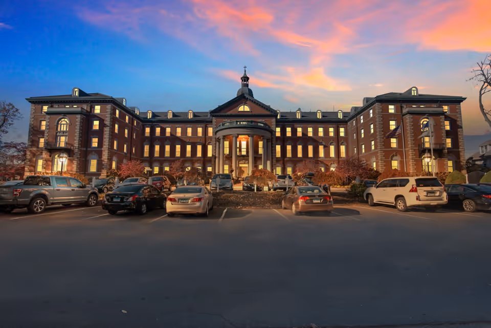A large, stately brick building with many lit windows at dusk, featuring a central entrance with columns and a small tower on top. Several cars are parked in front of the building in a parking lot, and the sky is colorful with shades of blue, pink, and orange.
