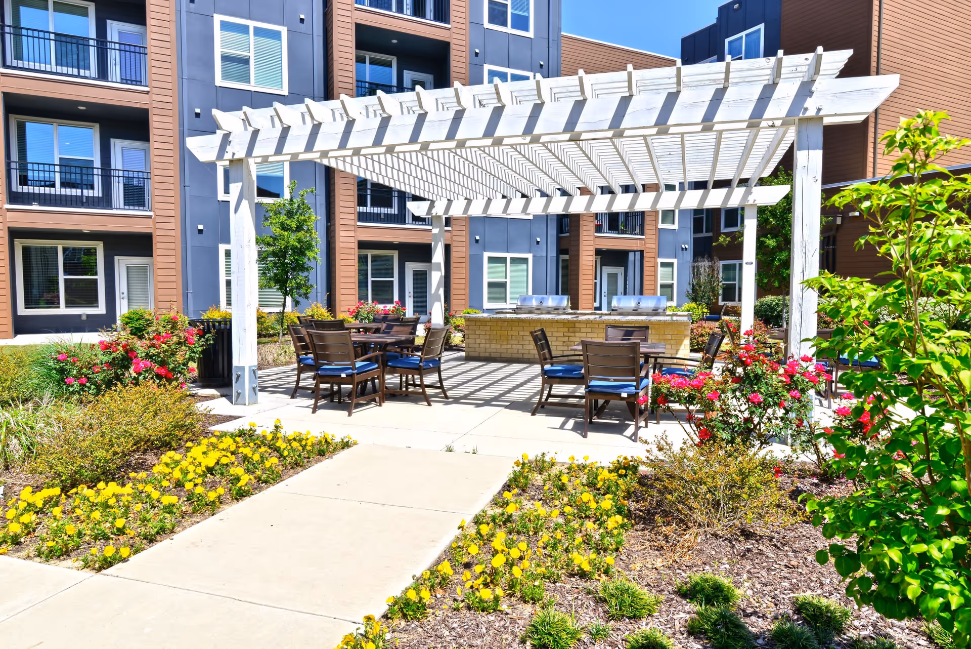 Outdoor patio area with a white pergola providing partial shade over several tables and chairs with blue cushions. The patio is surrounded by landscaped flower beds with yellow and red flowers, and there are modern apartment buildings in the background.
