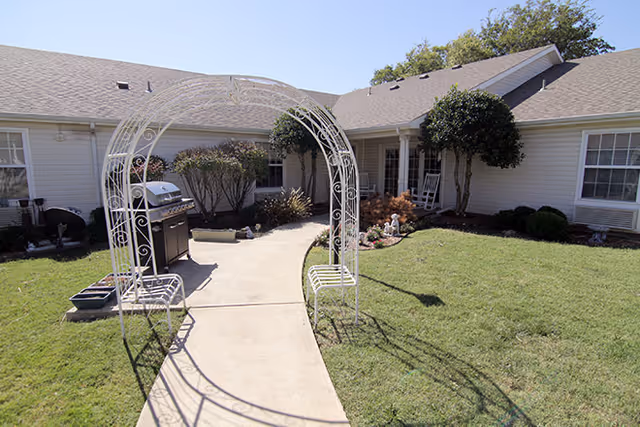 Outdoor courtyard area of an assisted living facility with a concrete pathway leading through a white metal archway. The courtyard has green grass, bushes, and trees, with a barbecue grill on the left side and a white rocking chair on the porch of the building in the background.