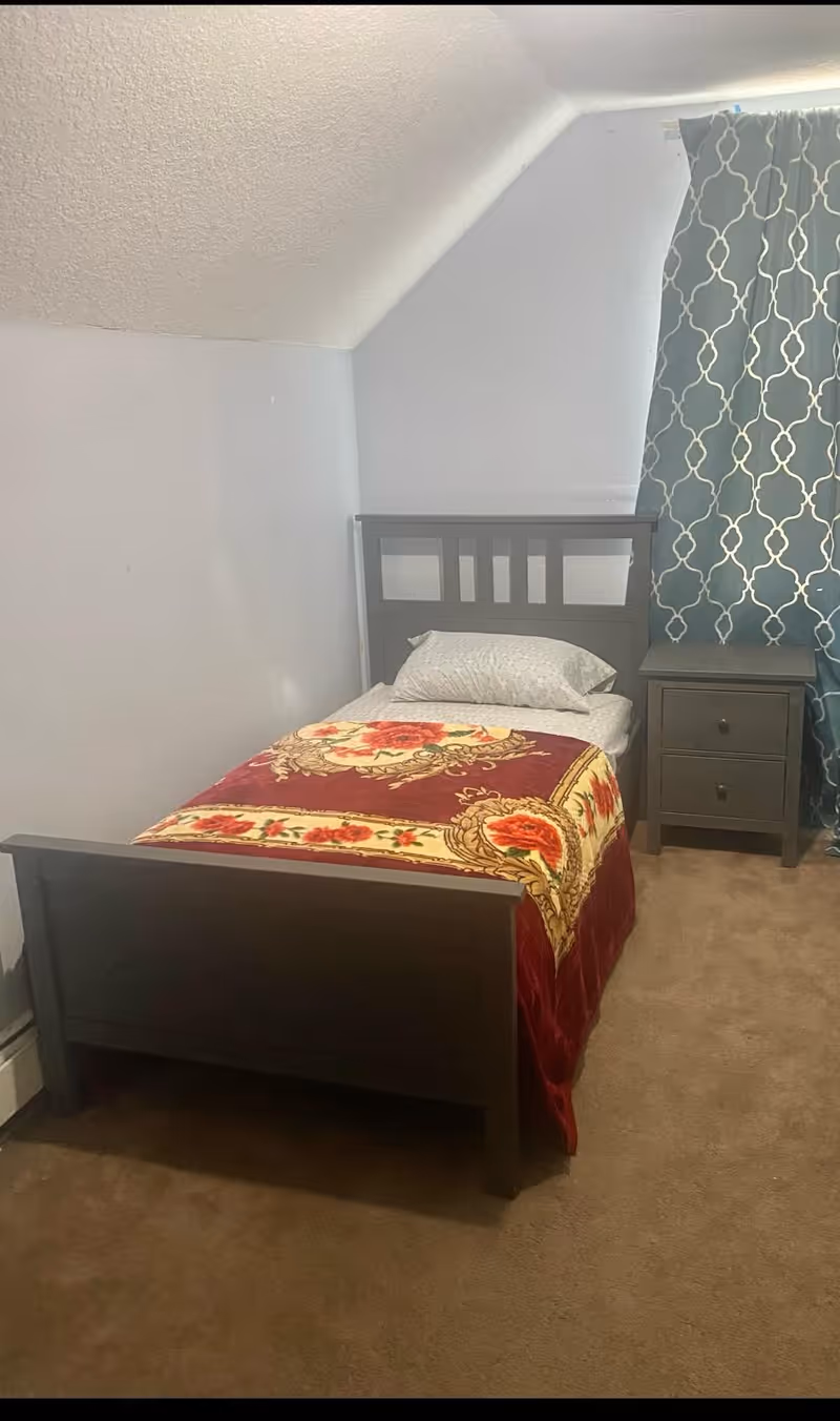 A small bedroom with a single dark wooden bed covered with a floral patterned blanket and a white pillow. Next to the bed is a matching dark wooden nightstand with two drawers. A window with blue and white patterned curtains is visible behind the nightstand. The walls are light-colored and the floor is carpeted.
