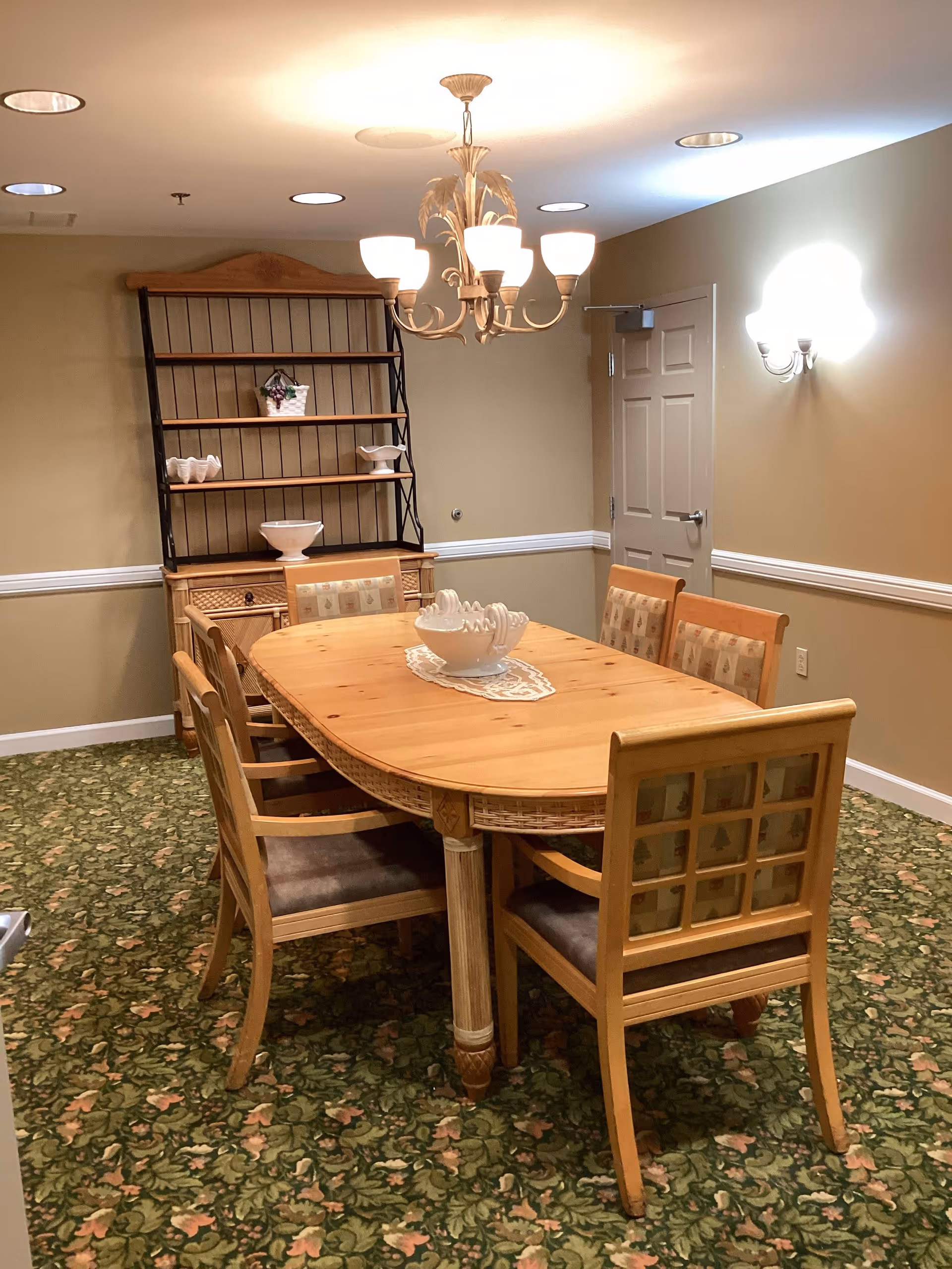 A dining room with a wooden oval table surrounded by six wooden chairs with cushioned seats. The floor is covered with a green floral carpet. A wooden hutch with shelves holding decorative bowls and a small basket is against the back wall. The walls are beige with white trim, and there is a chandelier with multiple lights hanging above the table. A closed door is visible on the right side of the room.