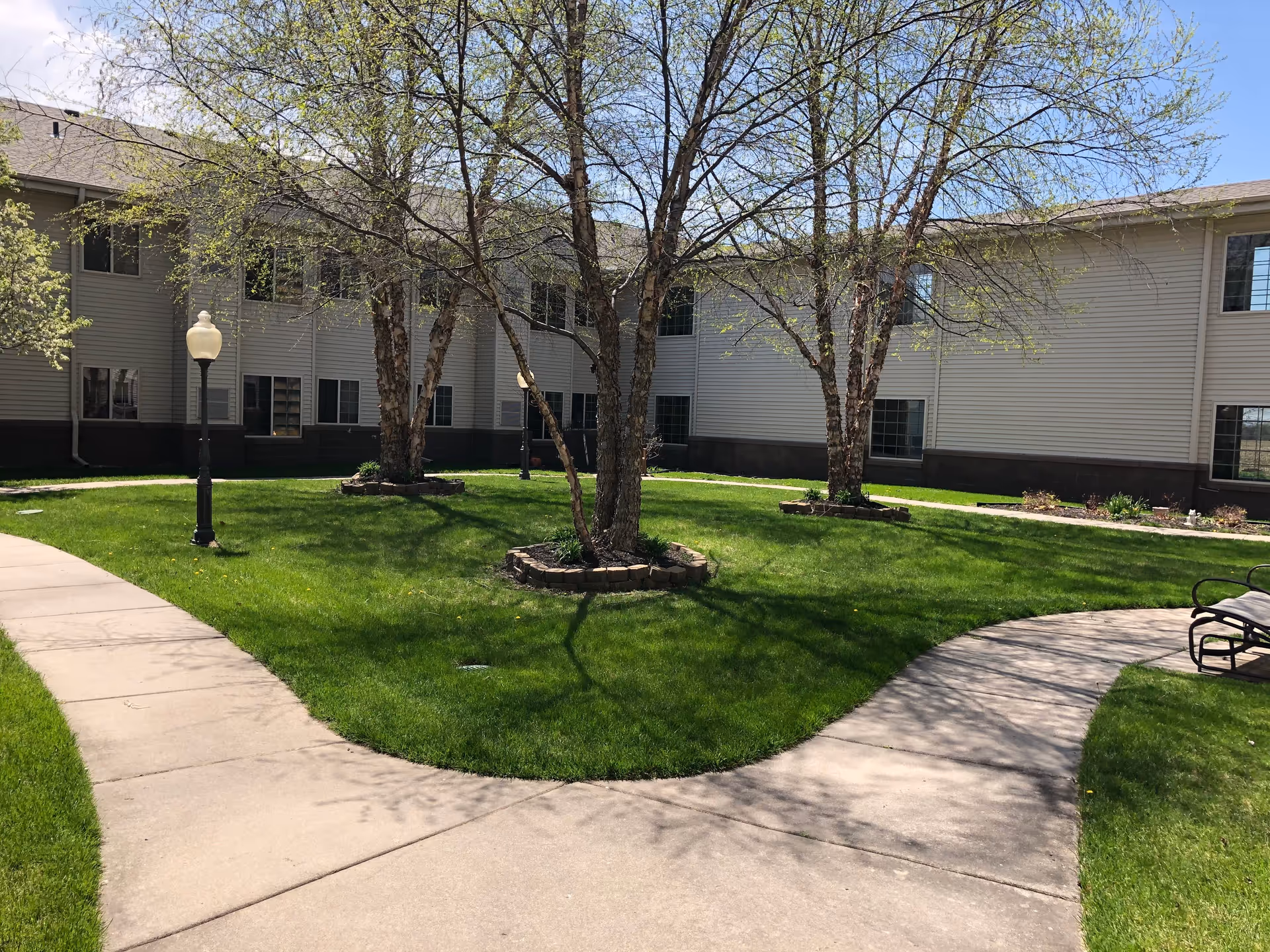 Outdoor courtyard area of Valley Lakes Assisted Living with green grass, three trees surrounded by small stone borders, a curved concrete walkway, a bench, and a two-story building with multiple windows in the background under a clear blue sky.
