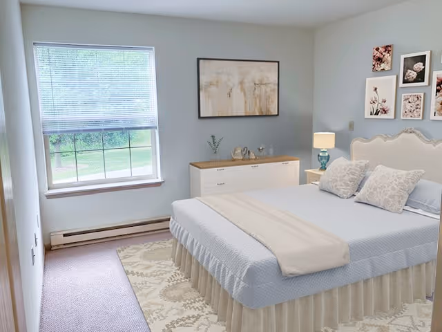 A bright and tidy bedroom with a large window letting in natural light. The room features a bed with a light blue bedspread, two patterned pillows, and a beige throw blanket. There is a white dresser with decorative items and a lamp on a nightstand next to the bed. The walls are decorated with framed floral artwork.