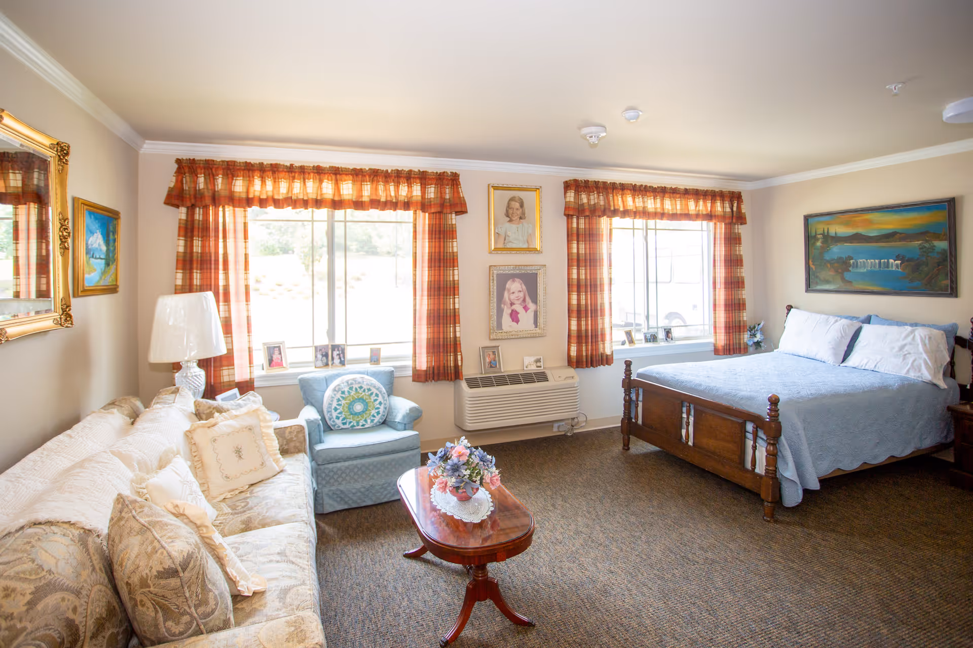 A cozy and well-lit assisted living room featuring a wooden bed with blue bedding, a beige patterned sofa with decorative pillows, a blue armchair with a round cushion, and a wooden coffee table with a floral centerpiece. The room has two large windows with red plaid curtains, framed portraits on the wall, a wall-mounted heater, and several framed paintings.