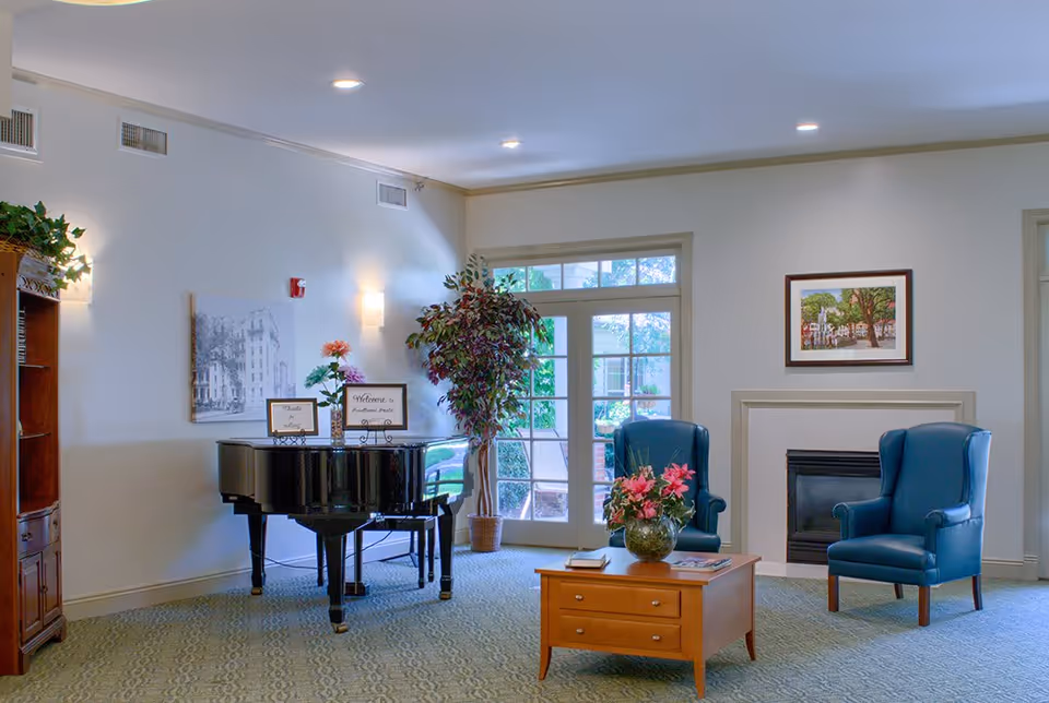 A bright and welcoming assisted living facility common area featuring a black grand piano with welcome signs on top, a potted plant next to a glass door leading outside, two blue armchairs near a fireplace, and a wooden coffee table with a flower vase and books.