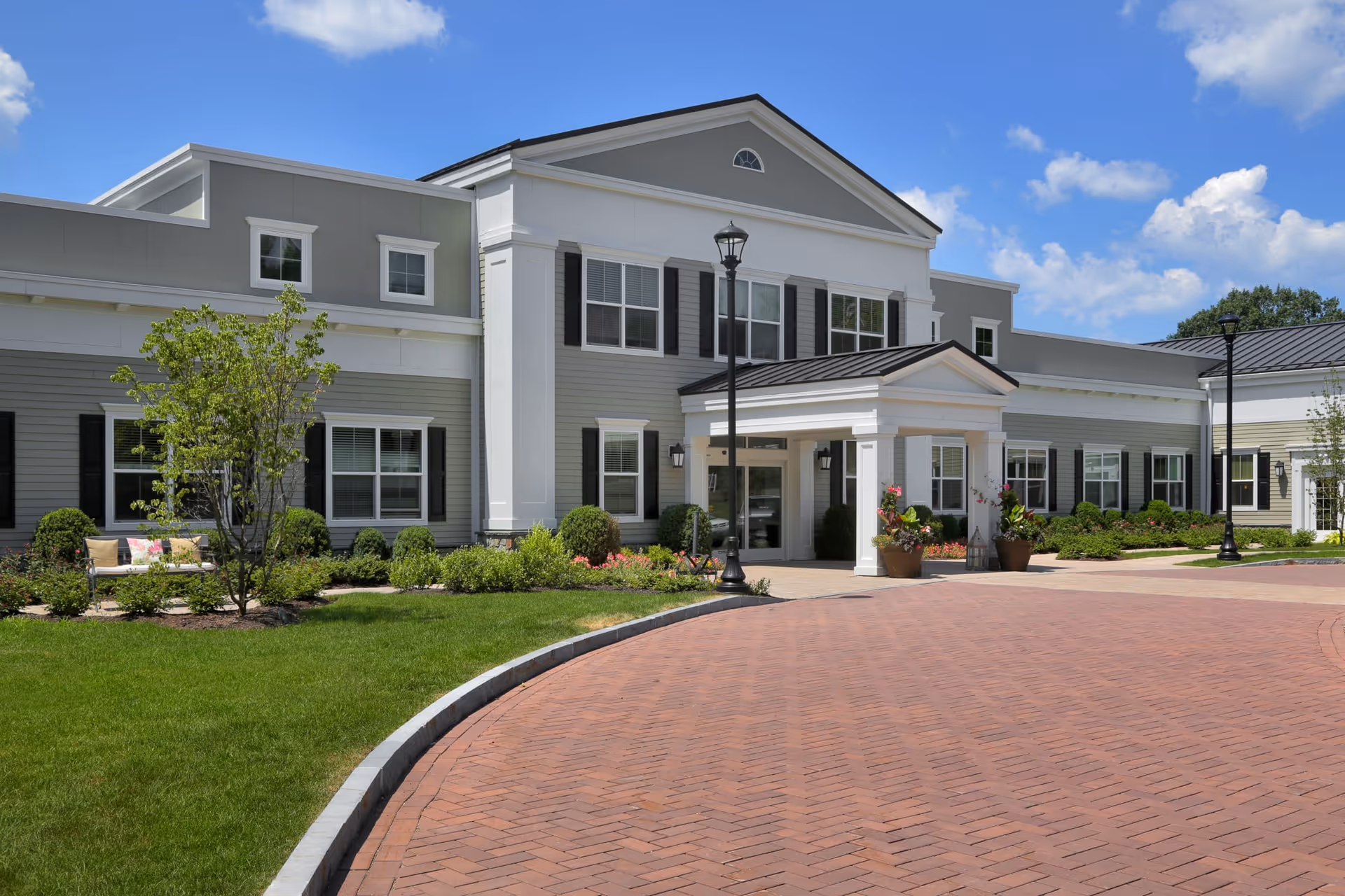 Exterior view of a senior living facility named Maplewood at Strawberry Hill, featuring a large two-story building with gray and white siding, black shutters, a covered entrance, landscaped greenery, and a brick driveway under a blue sky with some clouds.