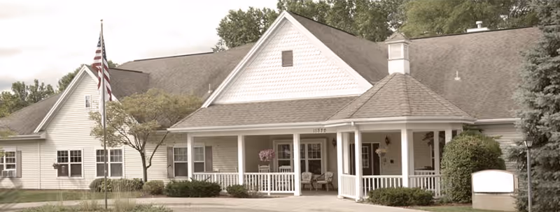 Front exterior of a beige senior living building with a covered porch, American flag, and surrounding landscaping.