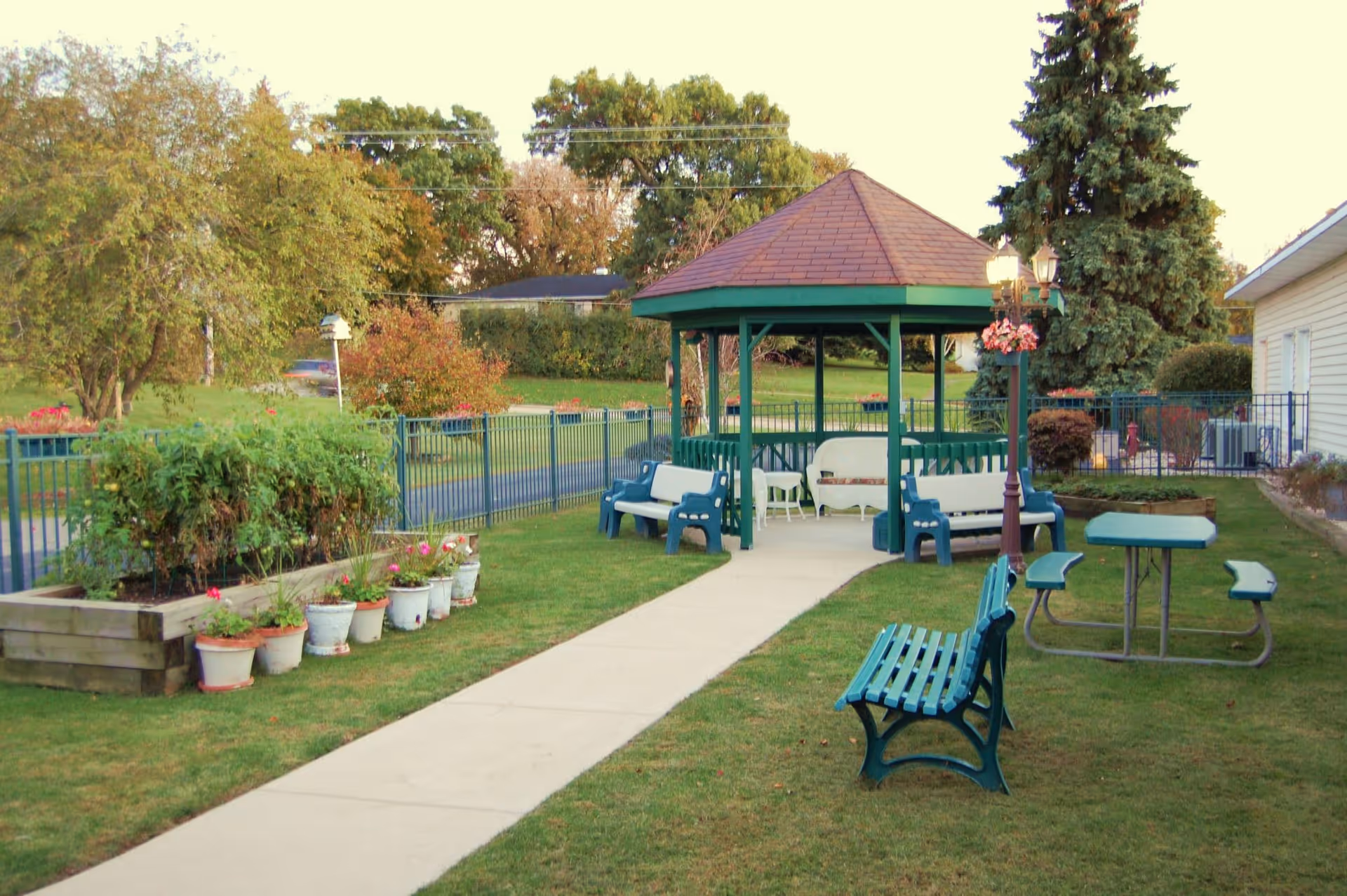 Outdoor garden area with a paved walkway leading to a green gazebo surrounded by benches. There are potted plants and a raised garden bed on the left side, a green bench and a picnic table with attached benches on the right side, and a lamppost with hanging flowers near the gazebo. Trees and a fence are visible in the background.