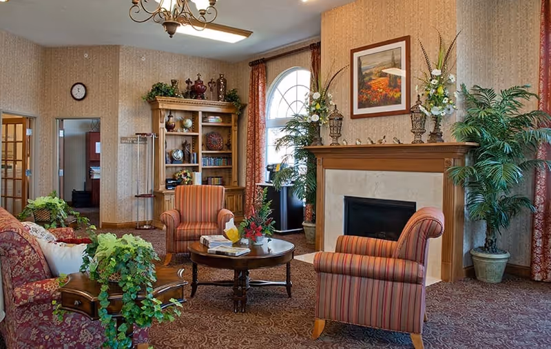 A cozy living room area in a senior living facility with patterned carpet and wallpaper. The room features a fireplace with decorative vases and flowers on the mantel, two striped armchairs, a round wooden coffee table with books and a flower arrangement, a floral upholstered sofa, and a wooden bookshelf filled with books and decorative items. Large windows with red curtains and several potted plants add to the warm ambiance.