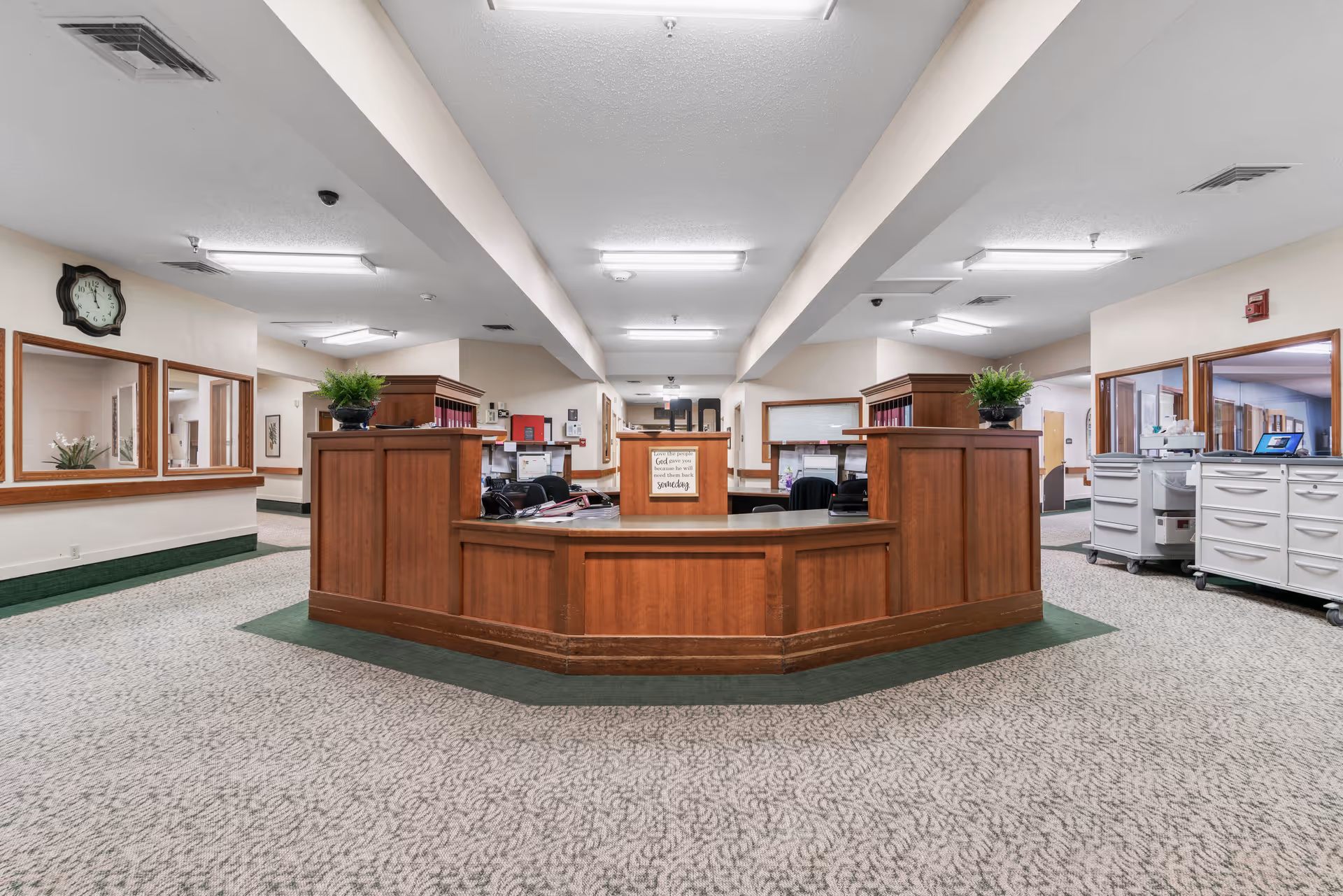 A spacious nursing facility reception area with a large wooden front desk in the center. The desk has plants on top and office supplies, with multiple windows and doors visible in the background. The carpet is patterned, and the ceiling has fluorescent lighting. There are filing cabinets and office equipment on the right side.