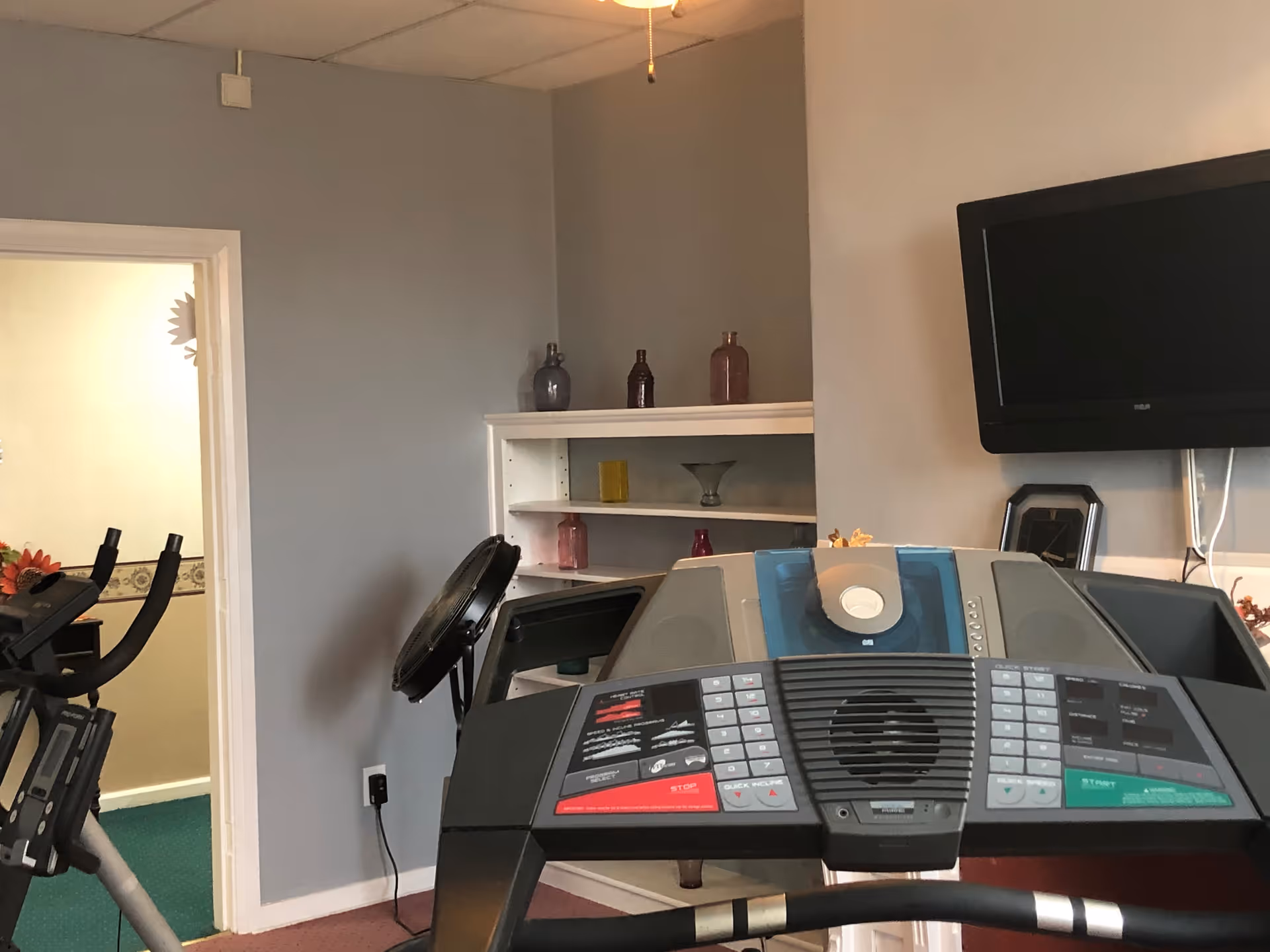 Indoor exercise room with a treadmill in the foreground, a stationary bike to the left, a wall-mounted TV on the right, and a white shelving unit with decorative bottles against a gray wall.
