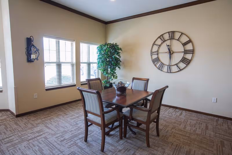 A small dining area with a wooden table and four cushioned chairs. The room has beige walls, a large decorative wall clock, a potted plant in the corner, and three windows with blinds letting in natural light.